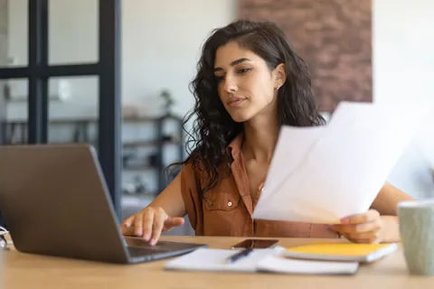 woman holding up two pieces of paper looking at her laptop