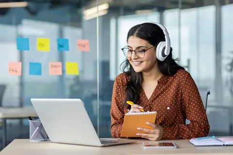 Smiling young woman with headphone on looking at her laptop and making notes