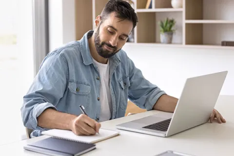 Man making notes on a notepad with a laptop in front of him